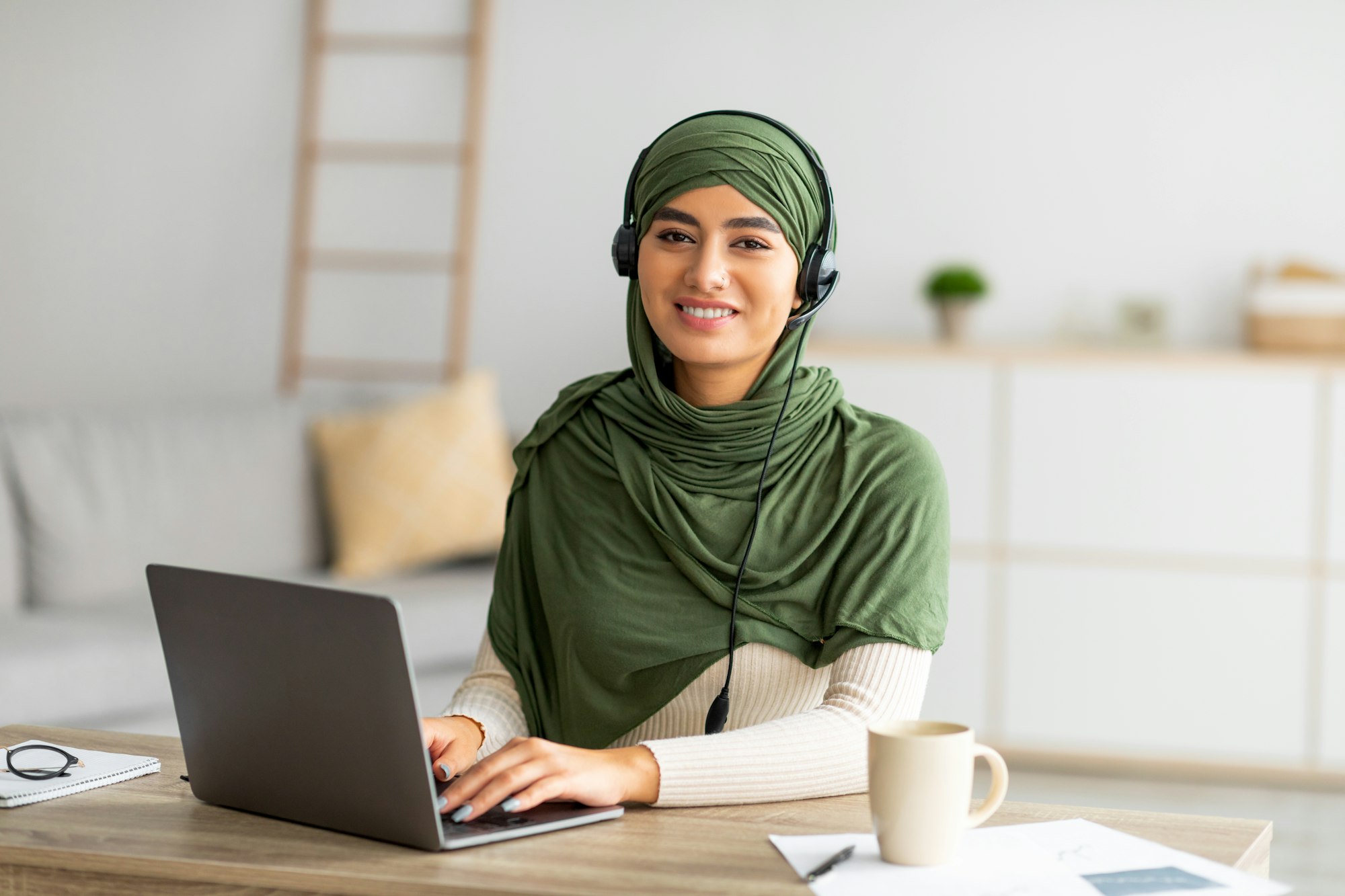 Young Arab female in hijab working on laptop from home, wearing headphones, using pc